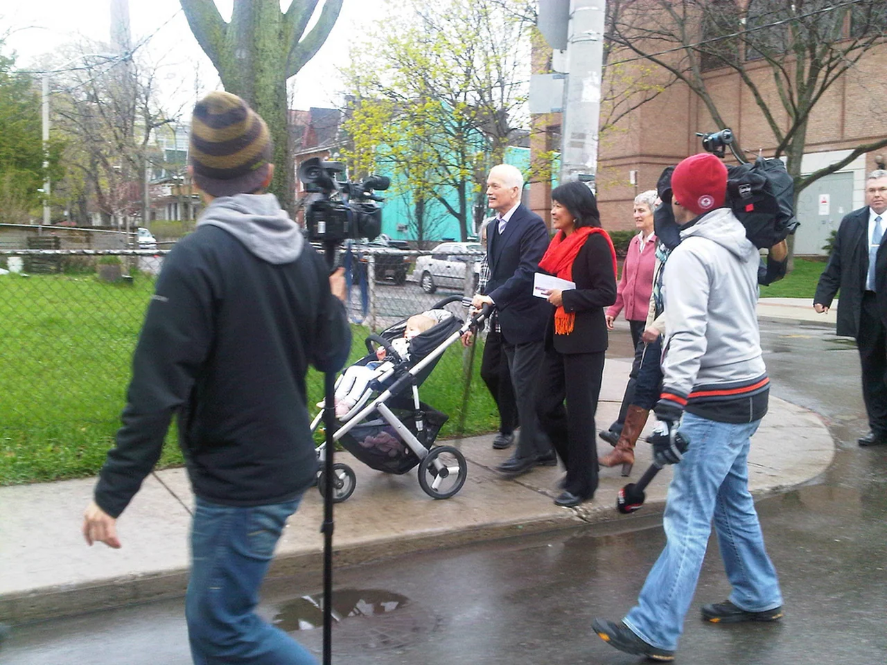 Olivia Chow, wearing red, walks beside Jack Layton on a Toronto sidewalk as cameras follow them.