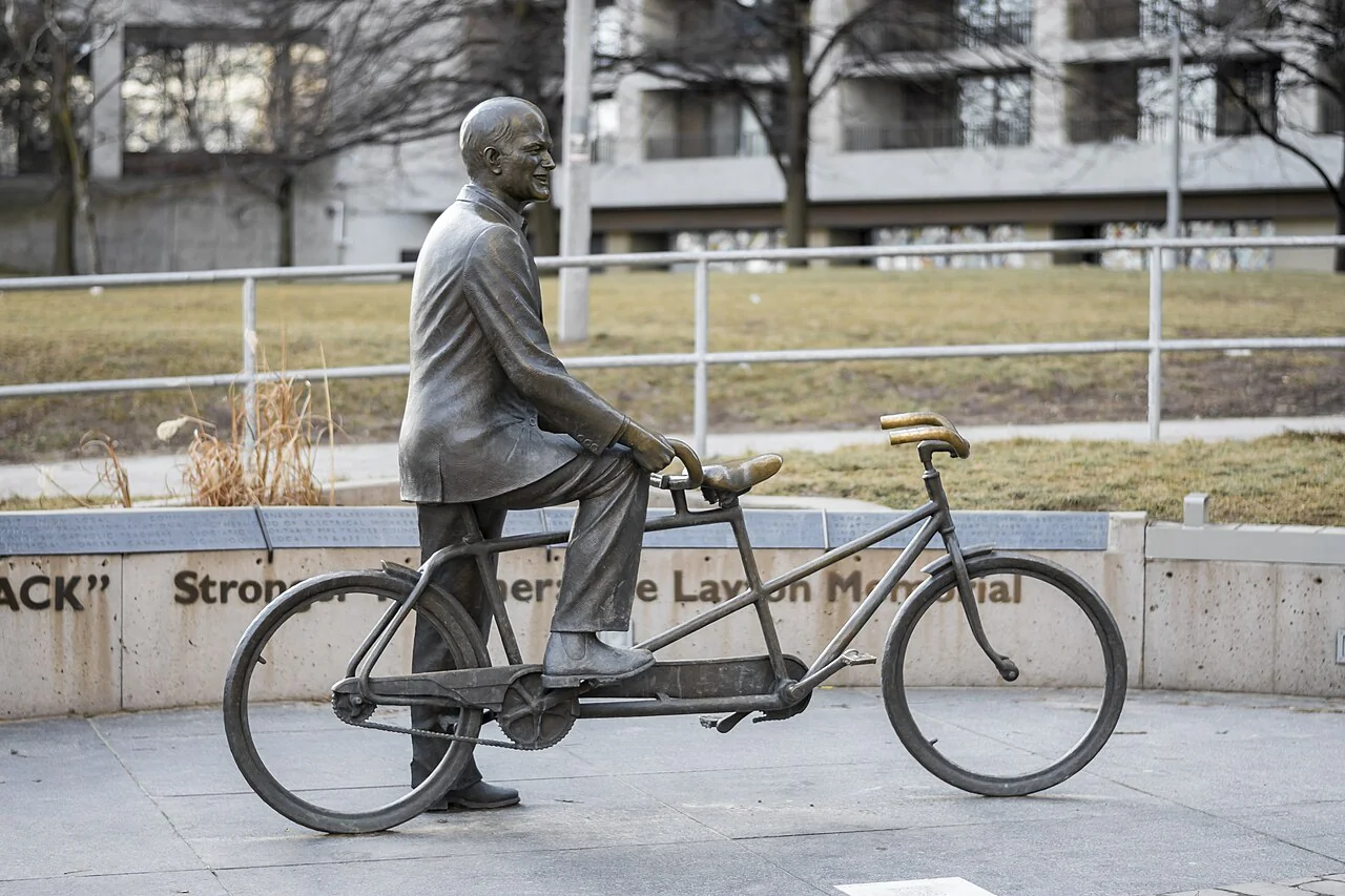 A bronze Jack Layton on the rear seat of a tandem bicycle, with the front seat empty.