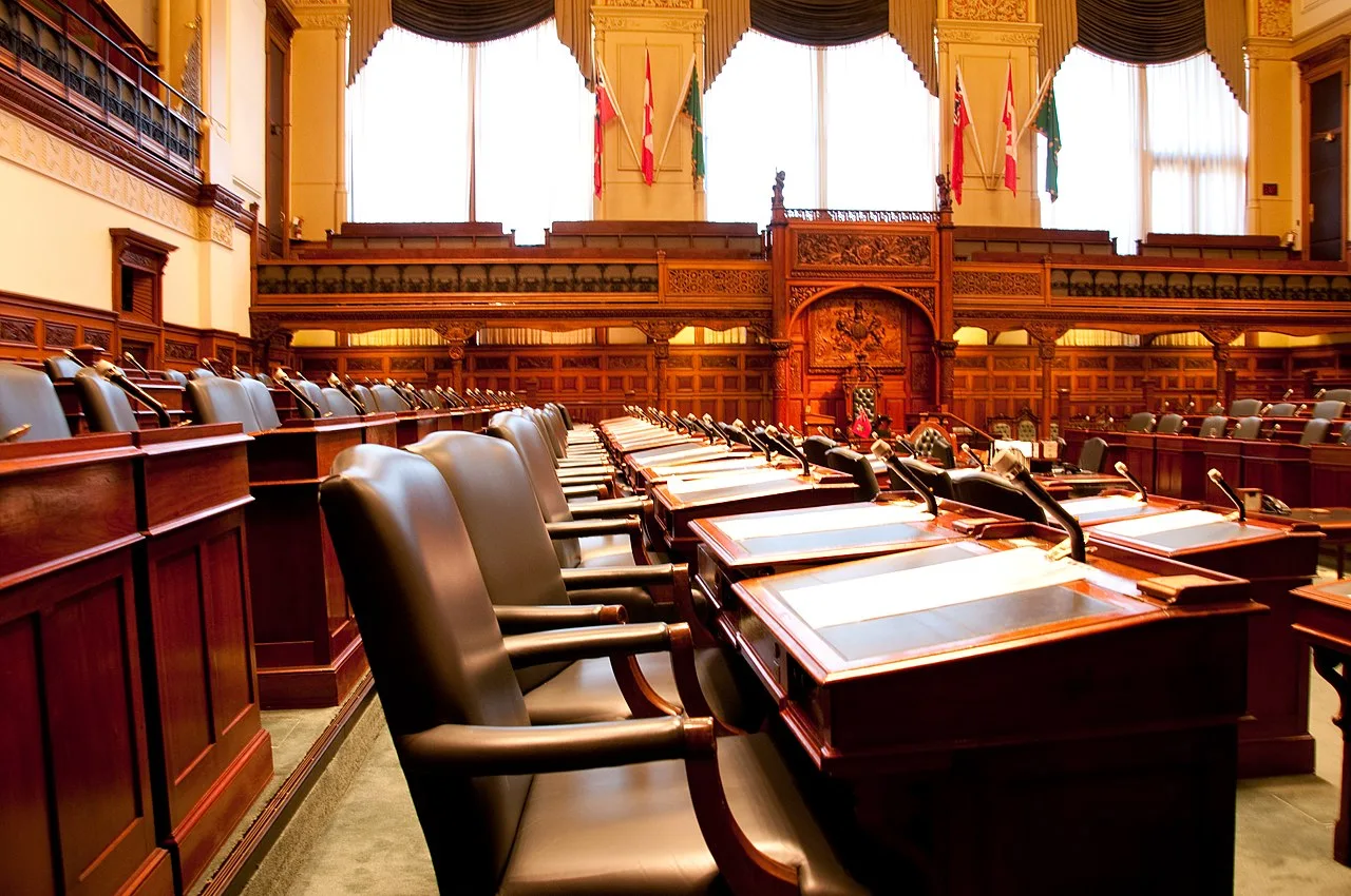 Rows of desks and the Speaker’s chair in the Ontario Legislative Chamber.