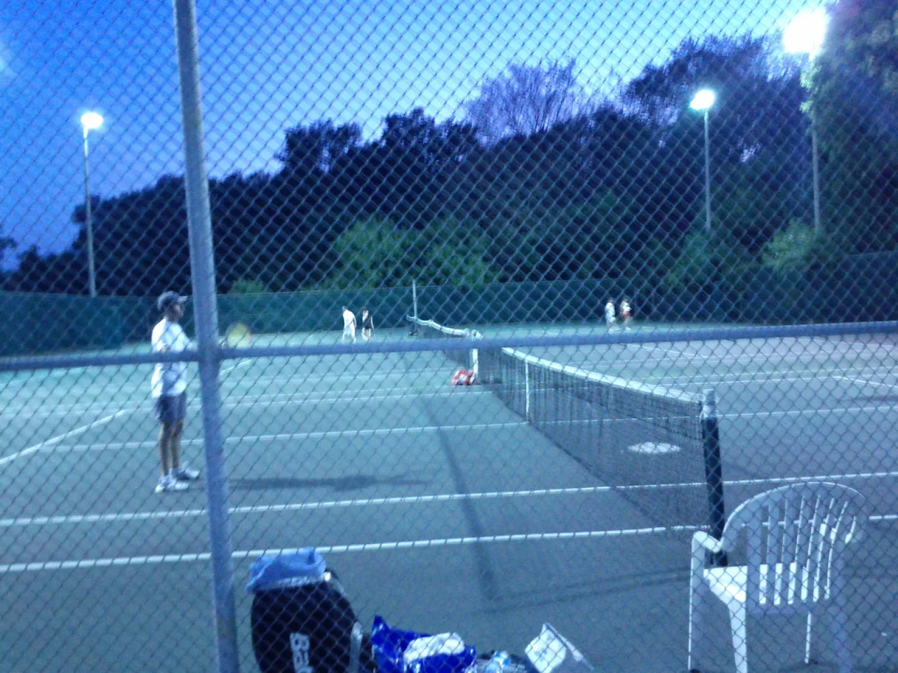 Players on the James Gardens tennis courts at dusk, viewed through the perimeter fence.