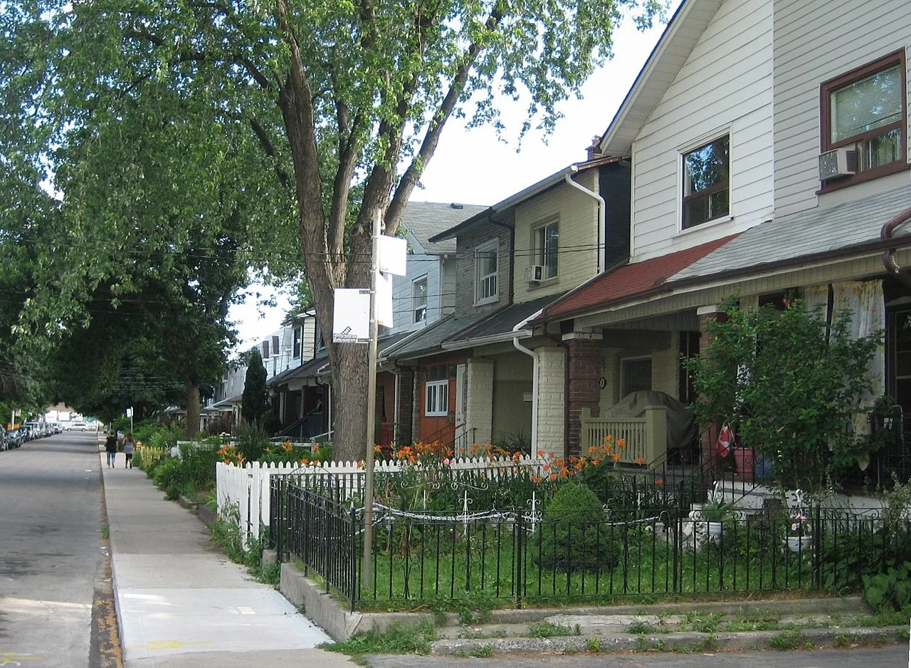 A row of houses with front porches and small gardens on a leafy Leslieville street.