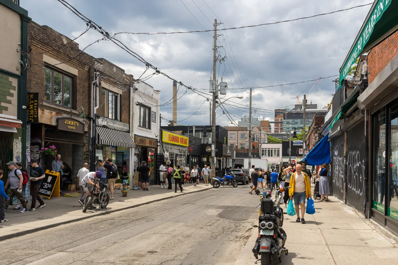People browse shops and walk along Baldwin Street in Toronto’s Kensington Market beneath overhead wires and a cloudy sky.